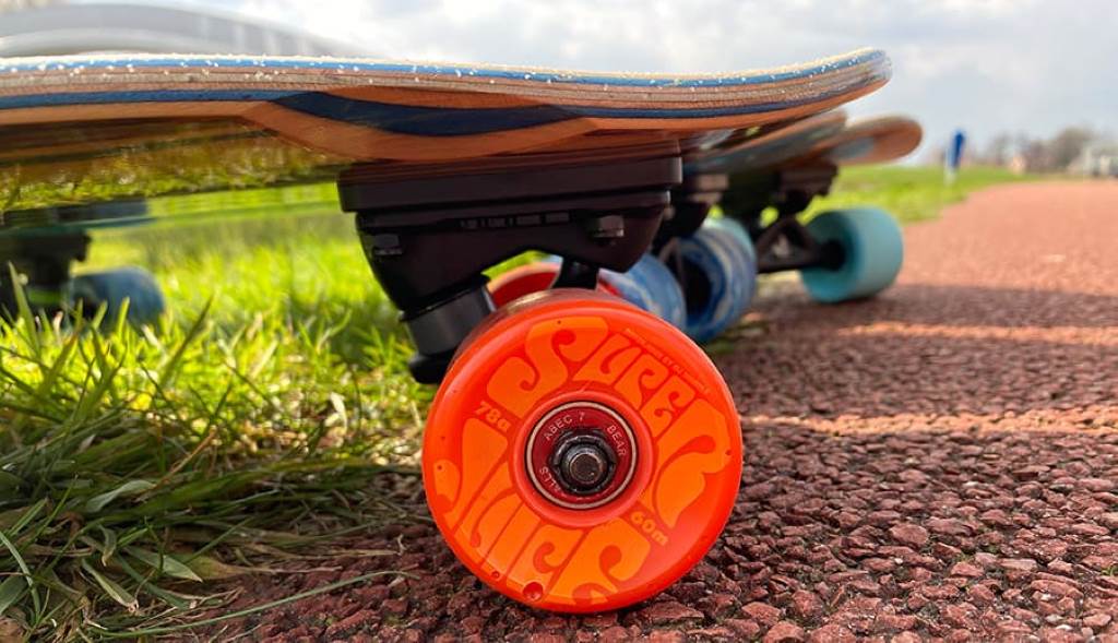 A close-up shot of a person's feet on a skateboard with bright orange cruiser wheels, rolling down a sunlit sidewalk.